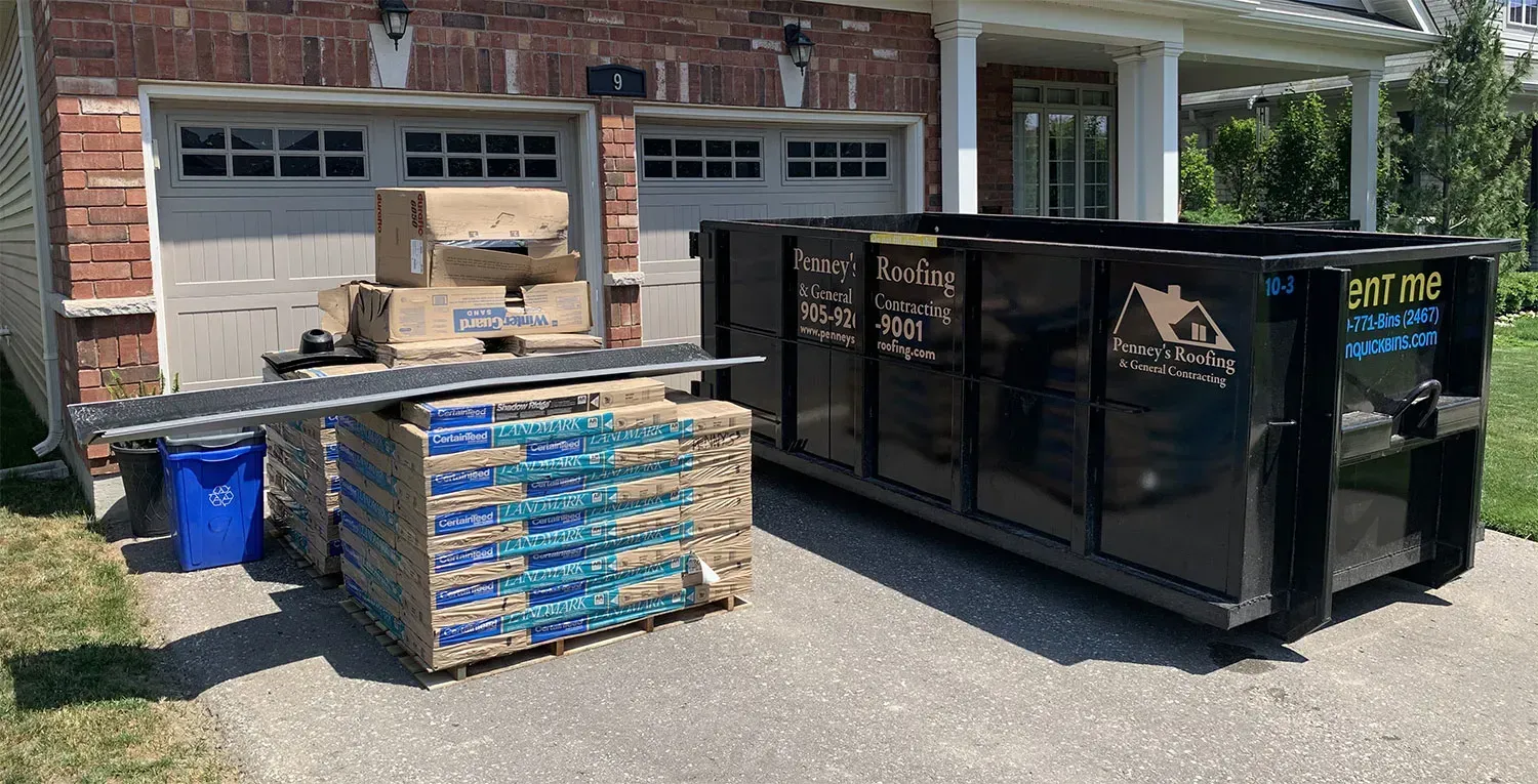 A black dumpster sits next to stacks of construction materials and boxes in front of a house.
