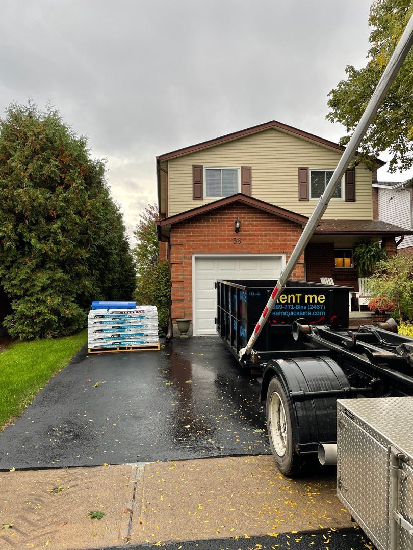 A dumpster truck in a driveway next to a house with pallets of building materials.