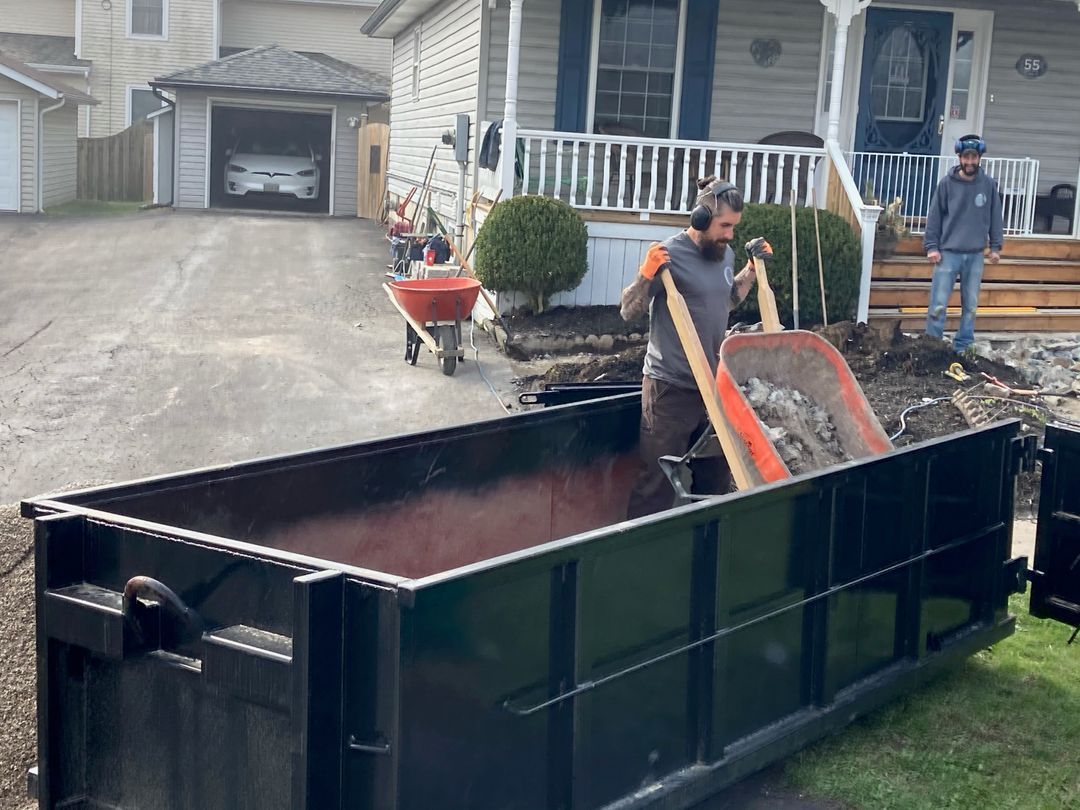 Men loading debris into a dumpster. One uses a shovel, another a wheelbarrow. House and driveway in background.
