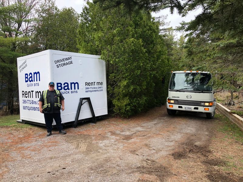 Man stands beside a white storage container, “bam” logo visible, with a small bus on a gravel road.