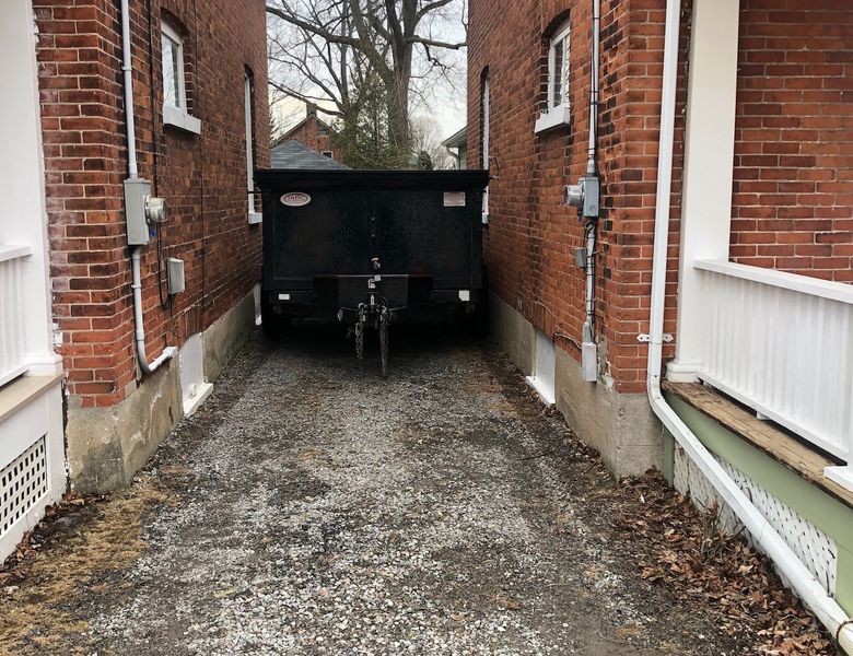 A black dumpster is parked in a narrow gravel driveway between two brick buildings.