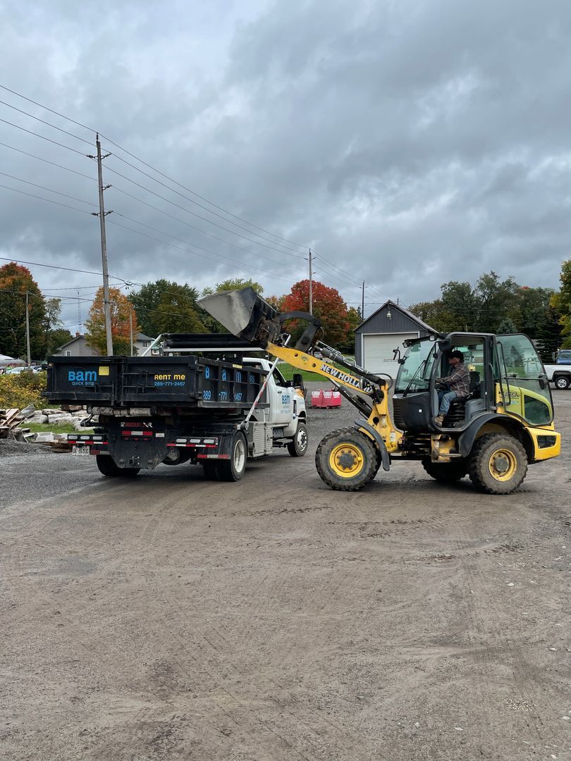 A yellow loader filling a black truck bed on a gravel surface under a cloudy sky.