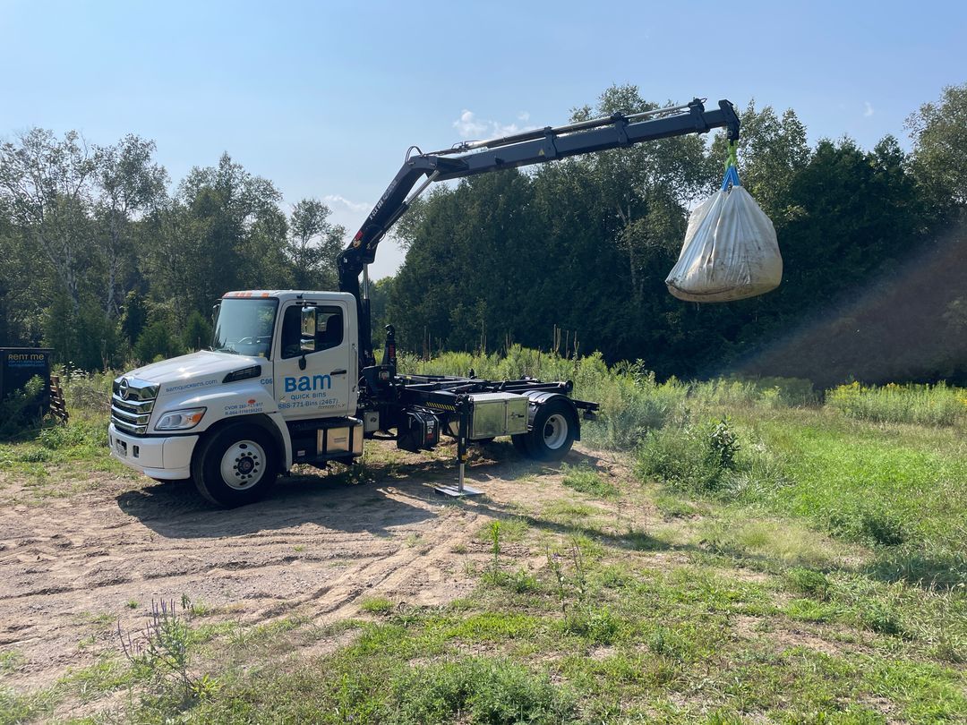 White truck with crane lifting a large white bag in a grassy field.