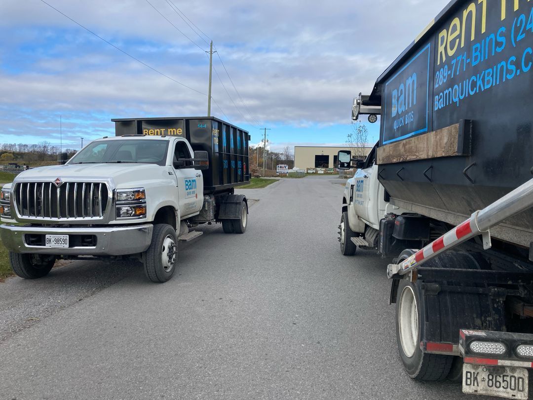 Two black dumpster trucks on a road, one with the dumpster raised. Blue sky, grey road.