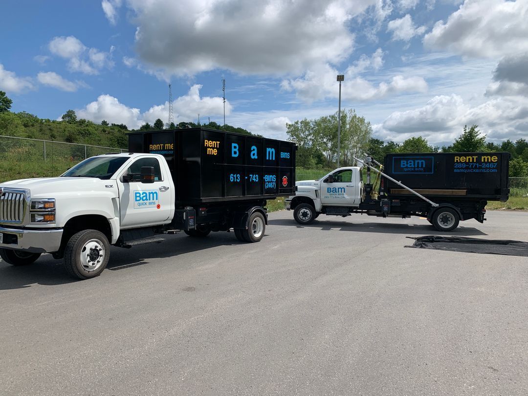 Two white dump trucks with black dumpsters in a parking lot on a sunny day.