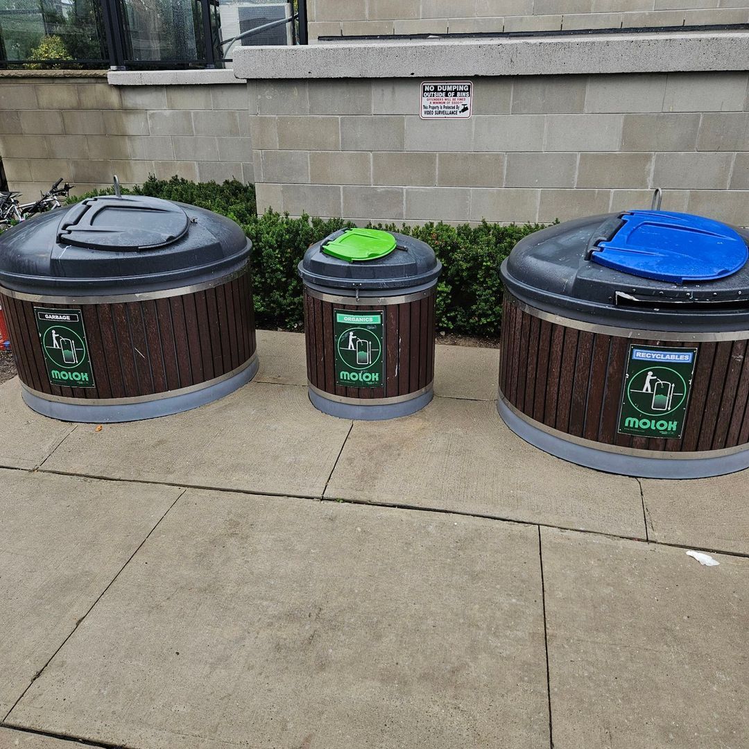 Three outdoor recycling bins with brown sides, different colored lids, on a sidewalk.
