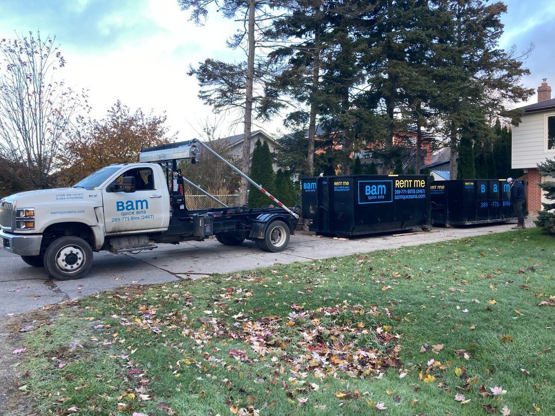 White truck pulling a black dumpster on a residential driveway. The sky is overcast, fall foliage.