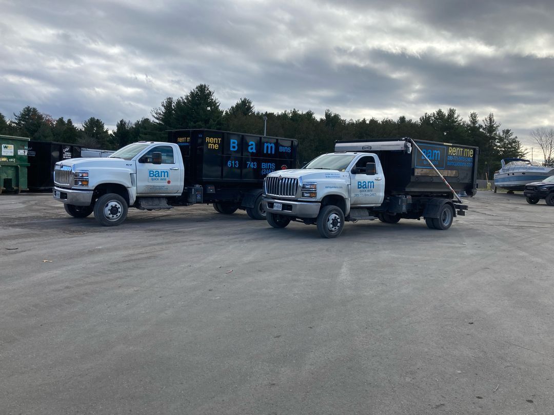 Two white dump trucks parked on gravel lot under cloudy sky. Trucks have 