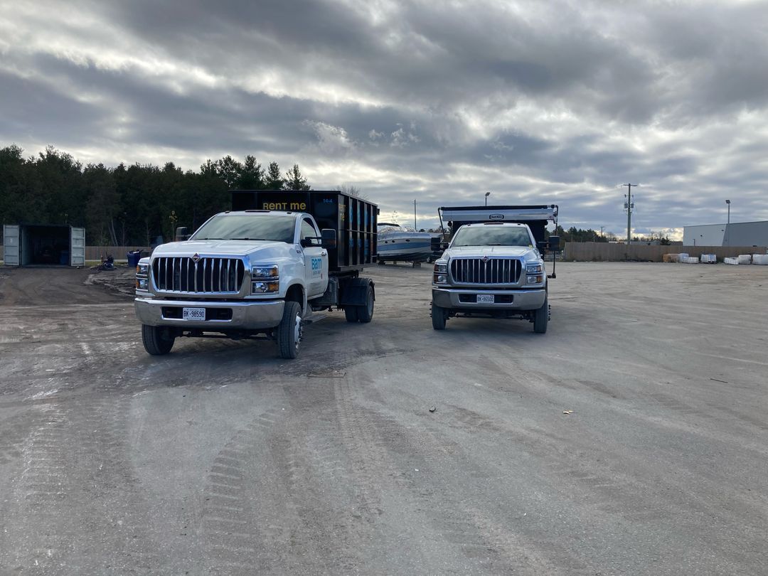 Two white dump trucks parked on gravel under a cloudy sky.