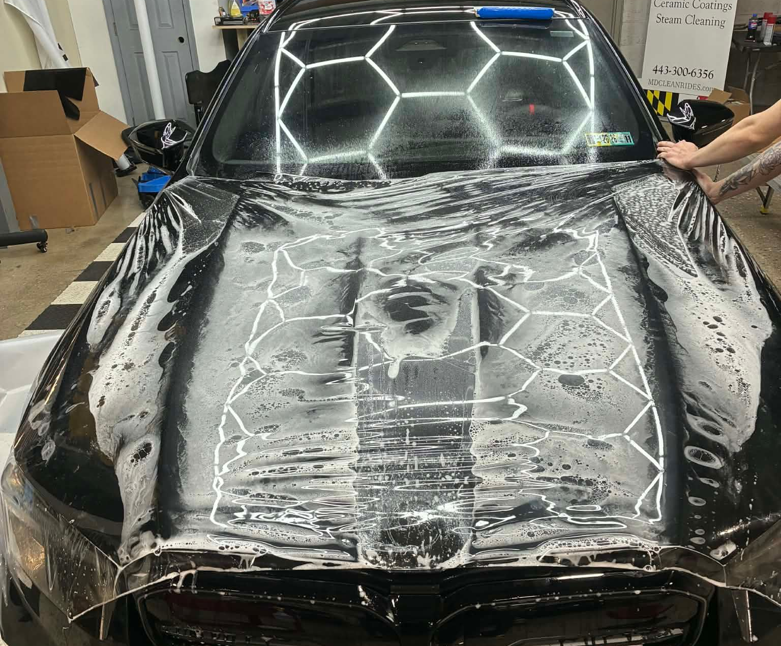 Black car hood covered in soapy foam during a wash, with bright lights reflected in the garage.