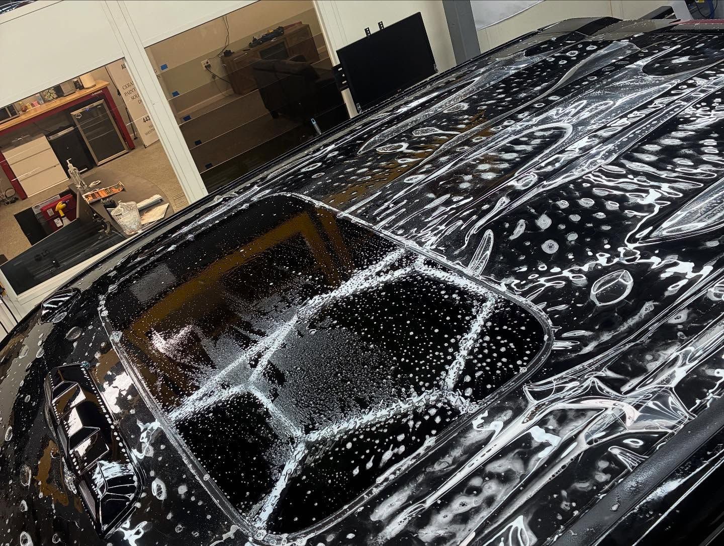 Black car hood covered in white soap suds and water droplets in a garage workshop