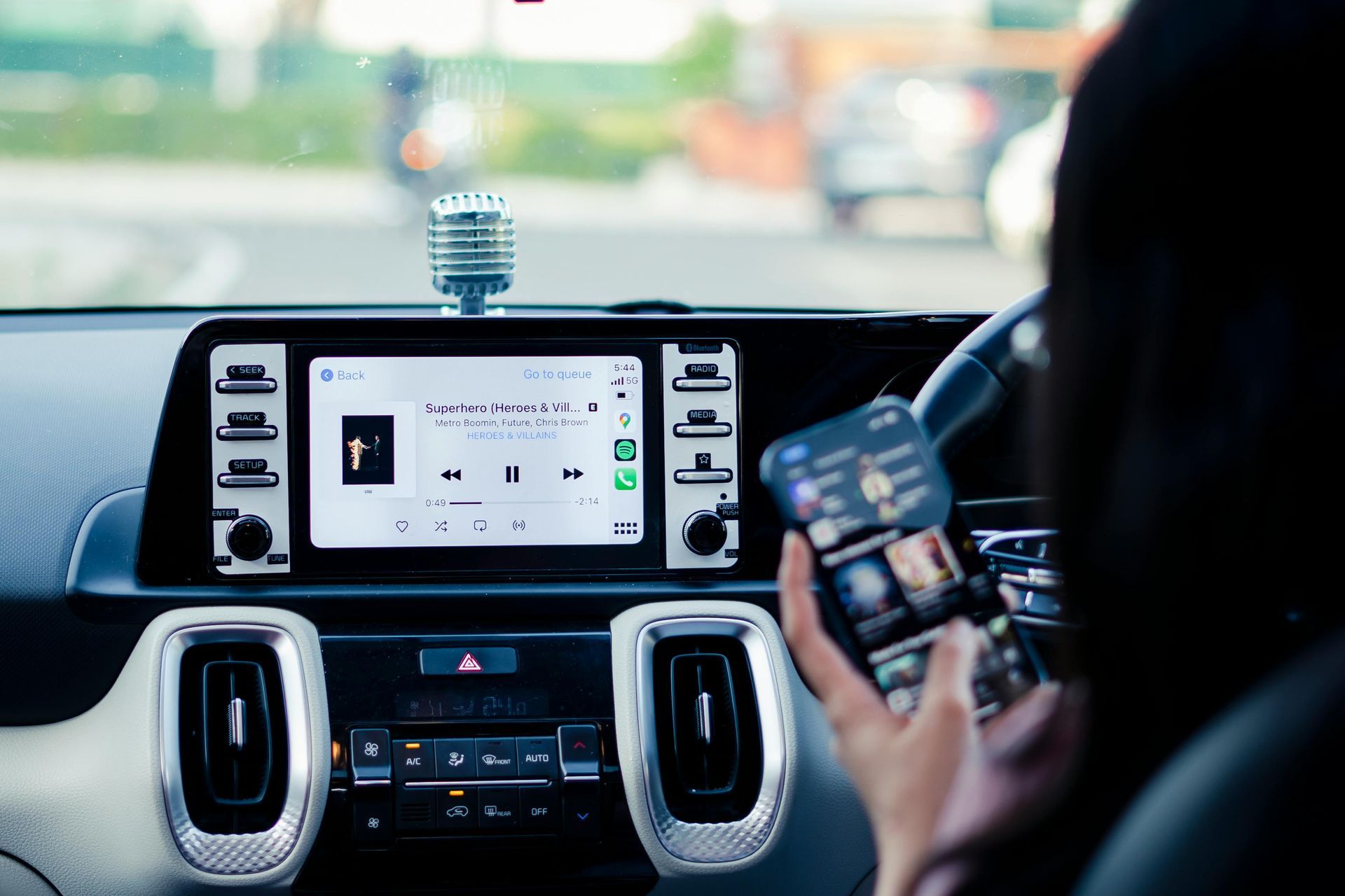 Person in car using a smartphone; car's dashboard shows media playing on its screen.