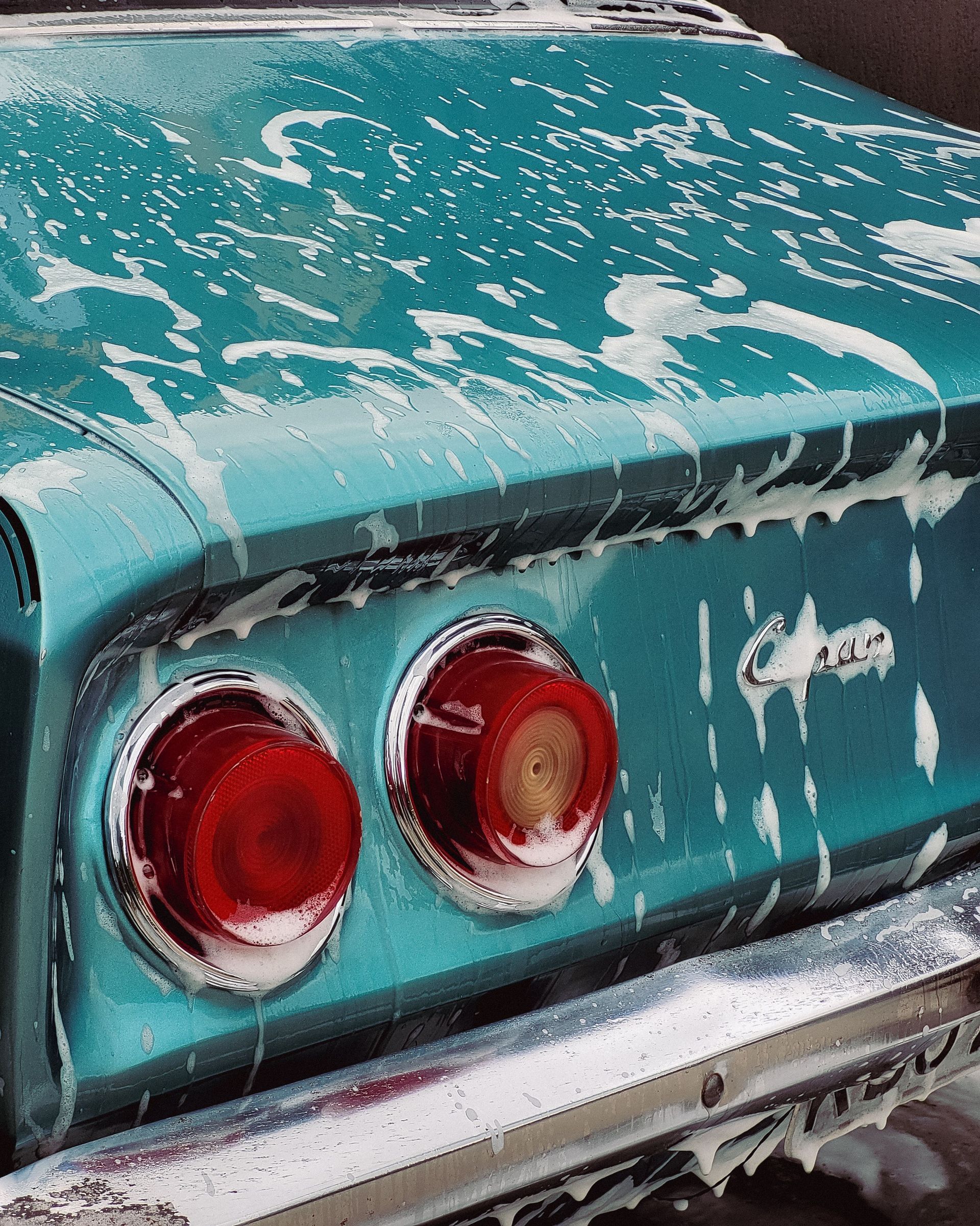 Rear of a teal classic car with red taillights, covered in streaks of white foam or paint splatter