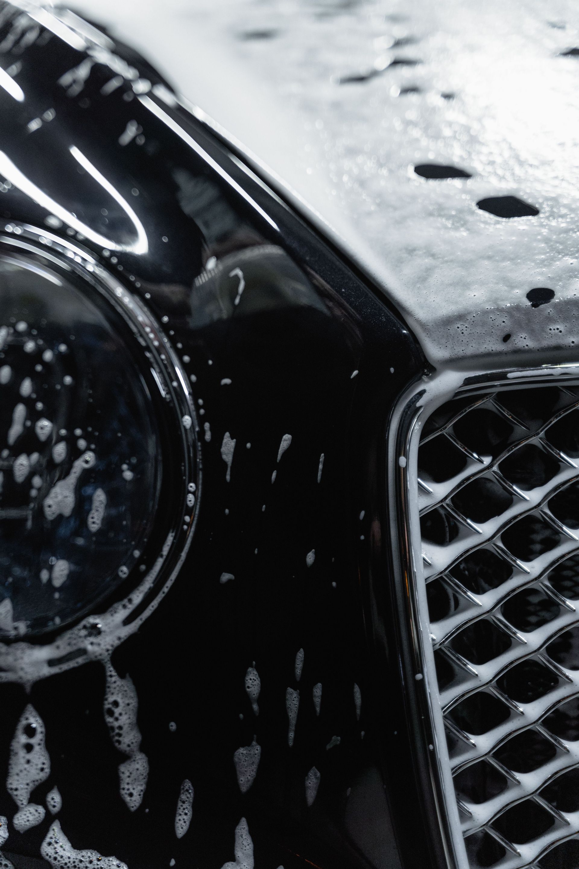 Close-up of a black car’s headlight and grille, covered in water droplets and soap suds