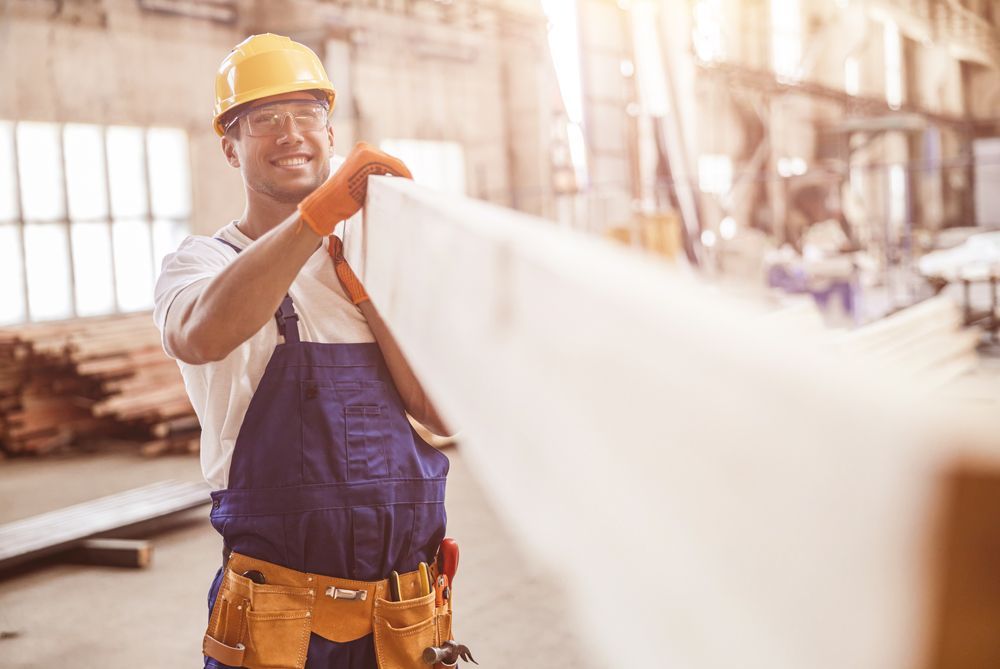 Construction worker holding a white wooden plank in a workshop, smiling.