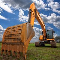 Yellow excavator with raised bucket on a grassy field under a cloudy blue sky
