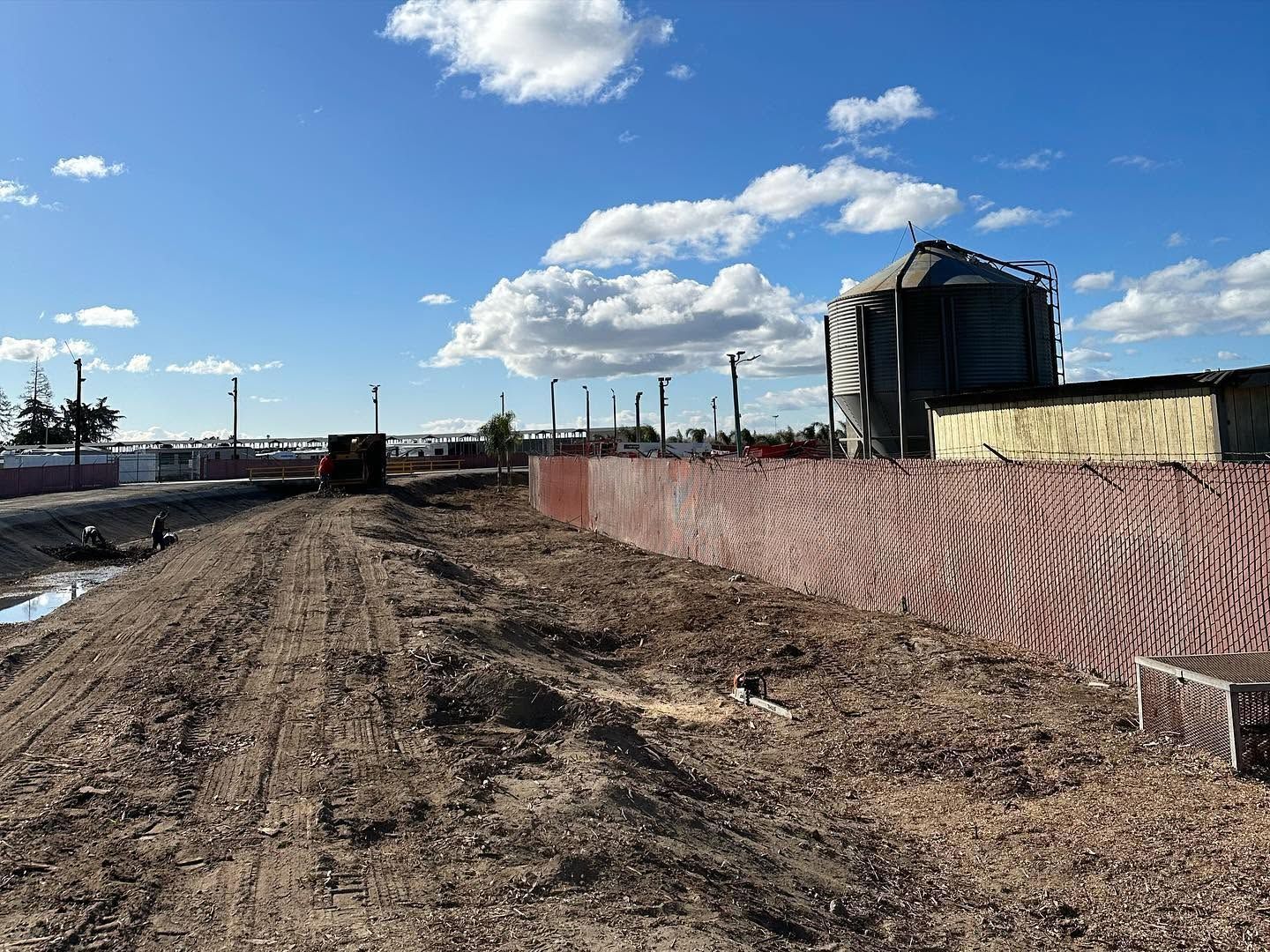 Dirt road beside a red fence and industrial tanks under a bright blue sky with clouds