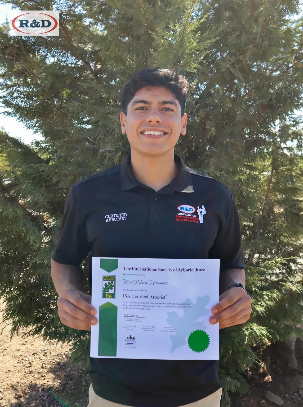 Person holding a certificate outdoors in front of trees, wearing a black shirt and smiling.