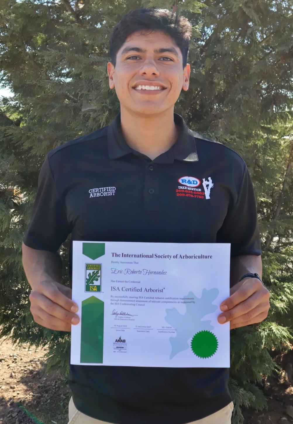 Young man outdoors smiling and holding a certificate in front of trees