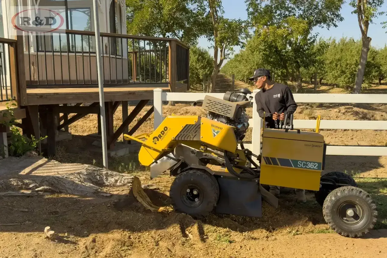 Yellow compact loader driving beside a wooden deck in a sunny yard, with a person operating it