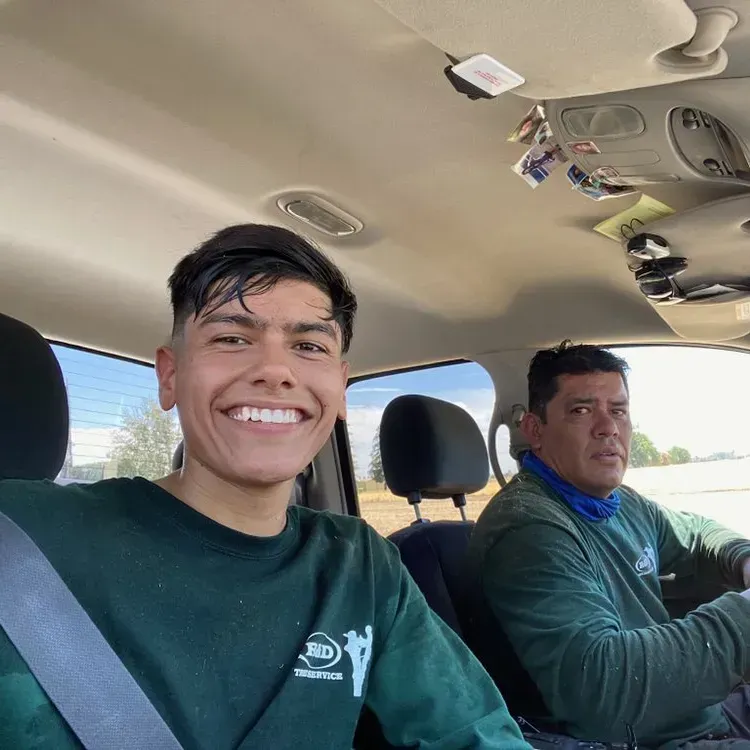 Two people in a car, one smiling in the front passenger seat and one driving, both in green shirts.