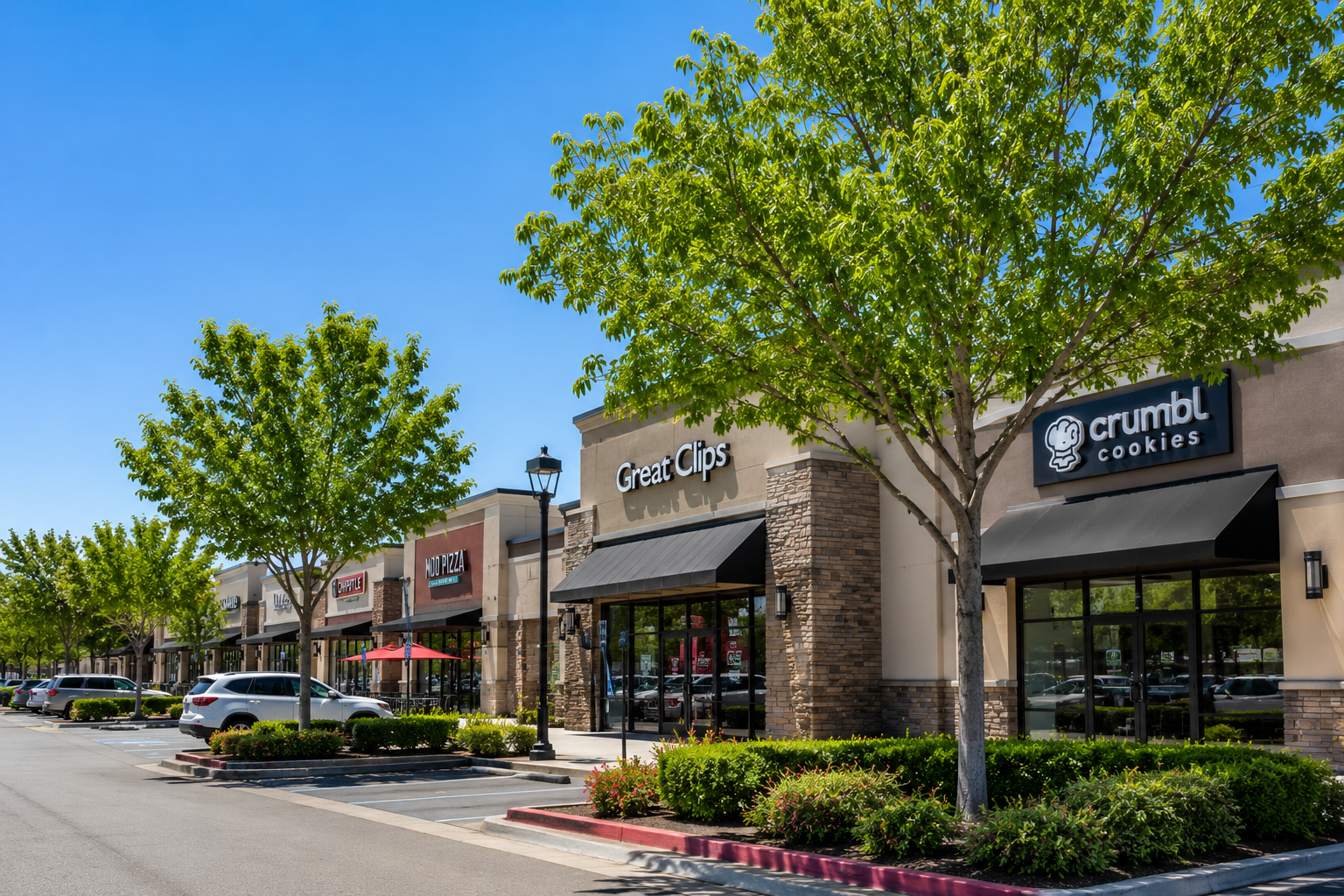 Shopping center storefronts with trees, parked cars, and a Crumbl Cookies sign on a sunny day