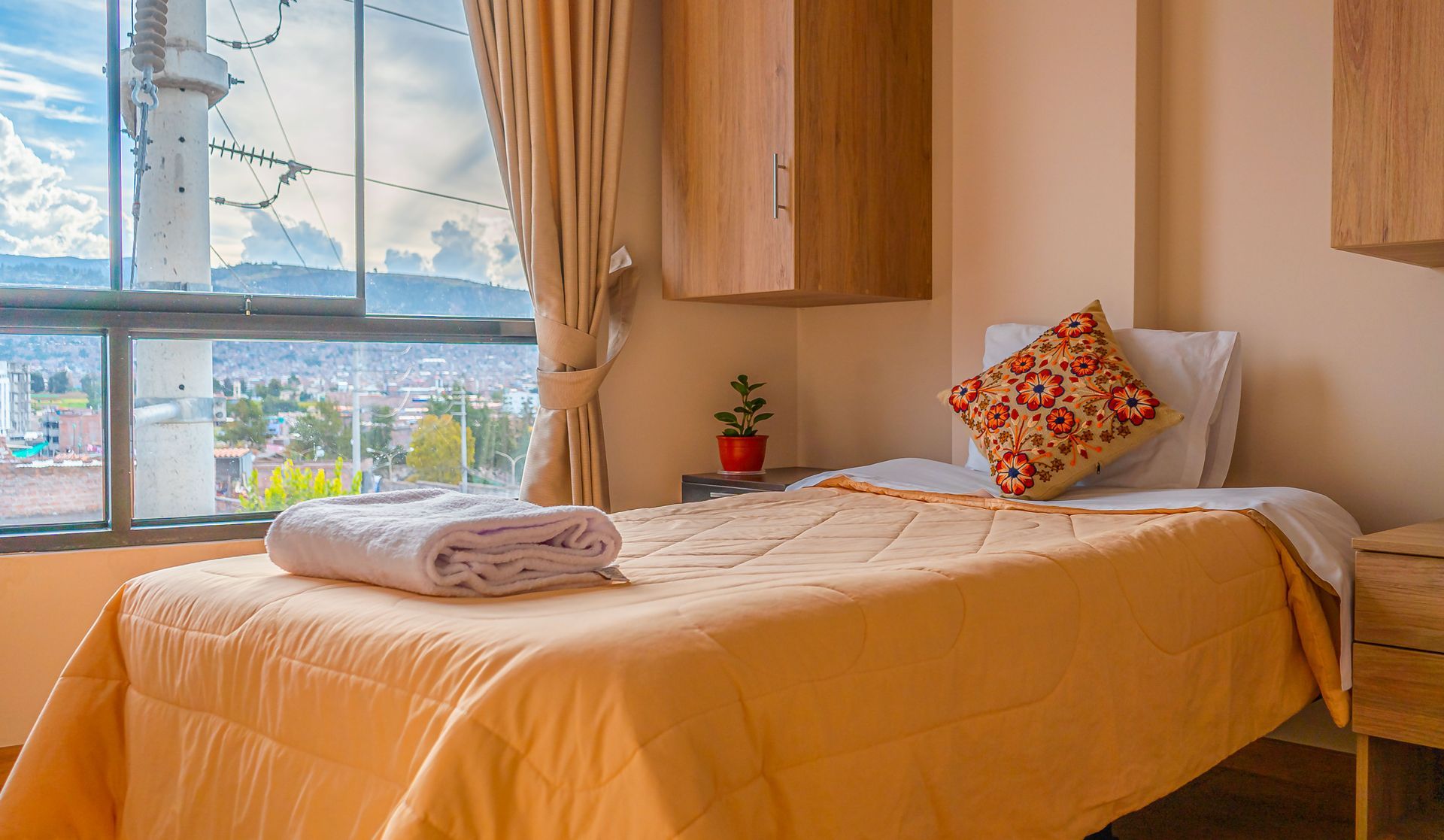 Sunlit bedroom with a neatly made bed, folded towels, floral pillow, and window view of city buildings
