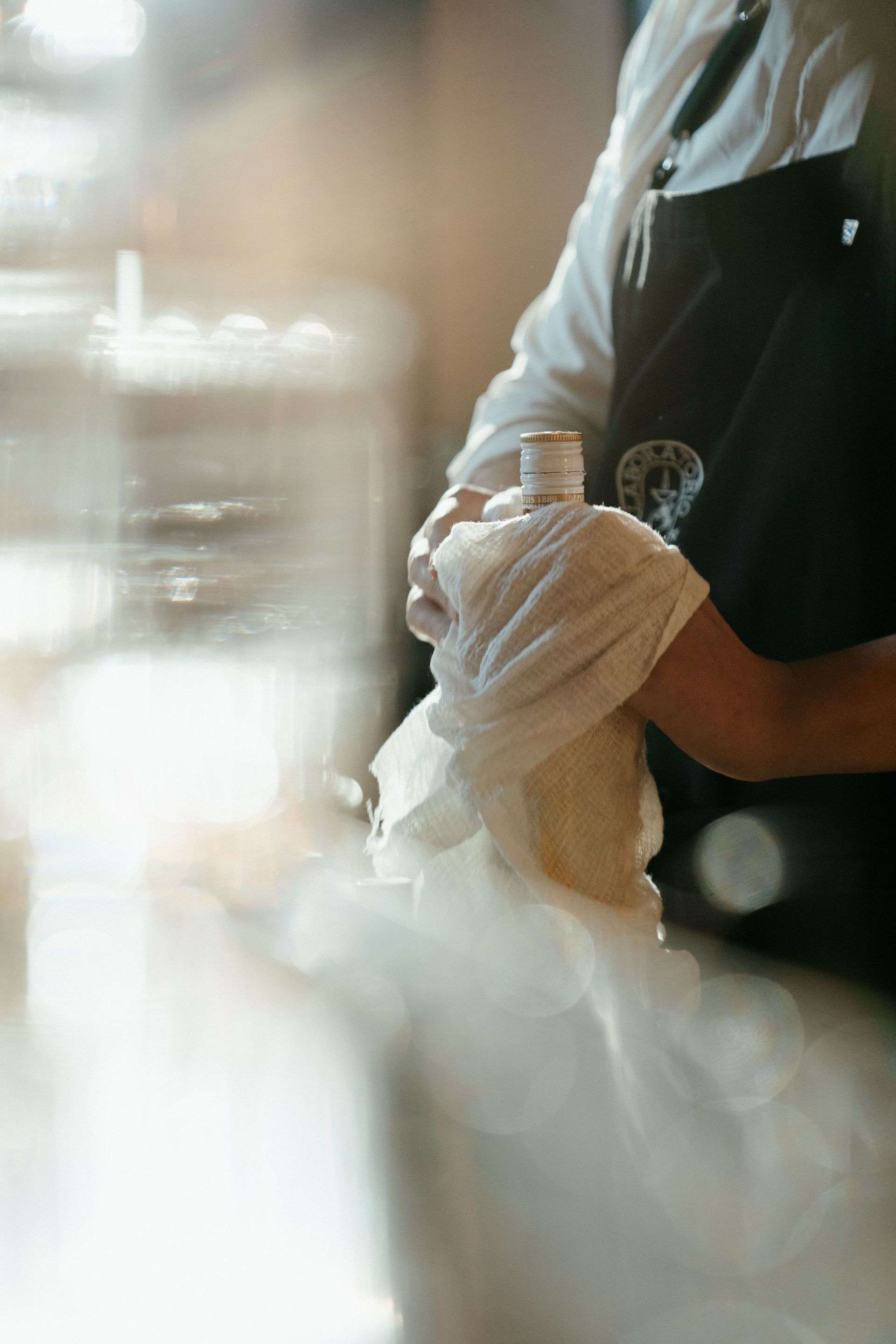 Person wiping a glass with a white towel behind a counter, softly lit in a café setting