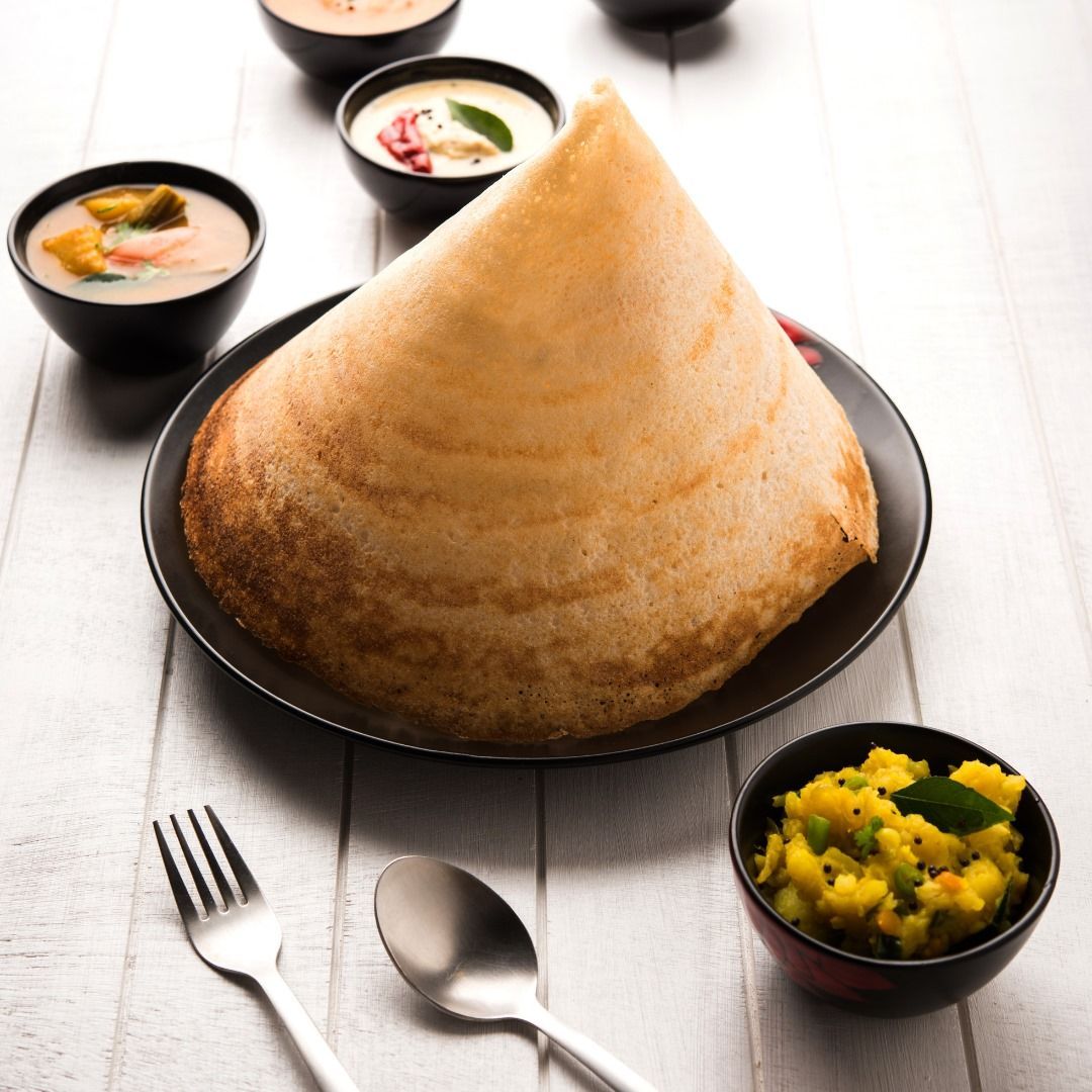 Cone-shaped dosa on a black plate with chutney and sambar bowls, along with a fork and spoon, on a white wooden table.