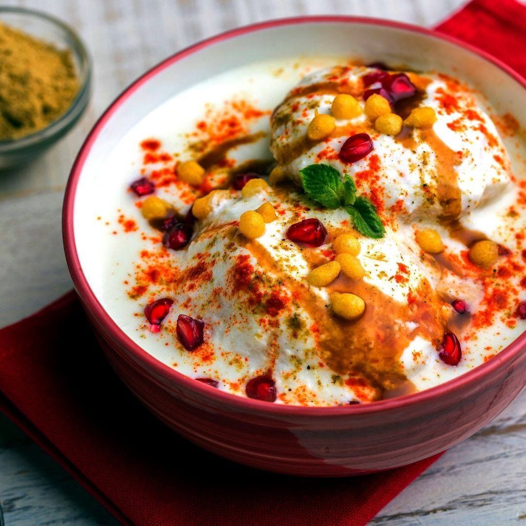 Dahi vada: yogurt-based dish with fried lentil fritters, topped with chutneys, spices, pomegranate seeds, and crispy boondi in a red bowl.