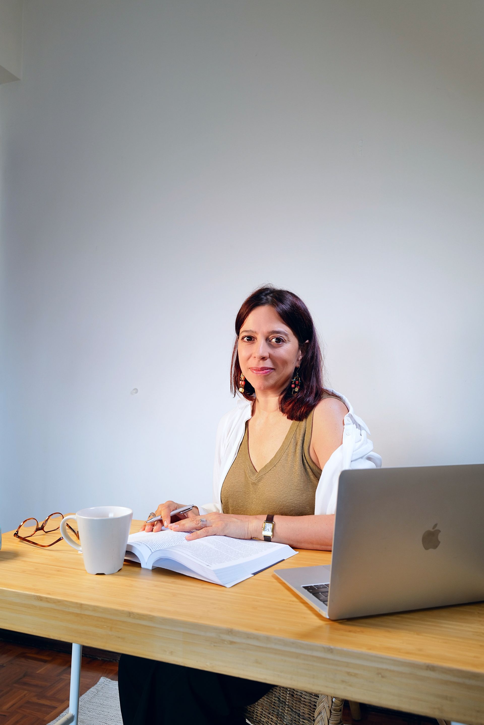 Mulher sentada à mesa com laptop, livro e caneca, sorrindo para a câmera.