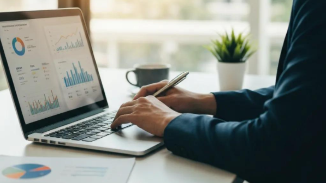 Person in suit works on laptop, viewing financial charts, in well-lit office.