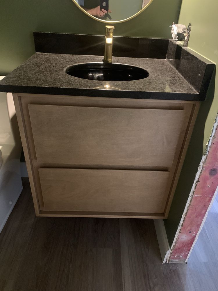 Small bathroom vanity with black countertop, round sink, and gold faucet beside a mirror.