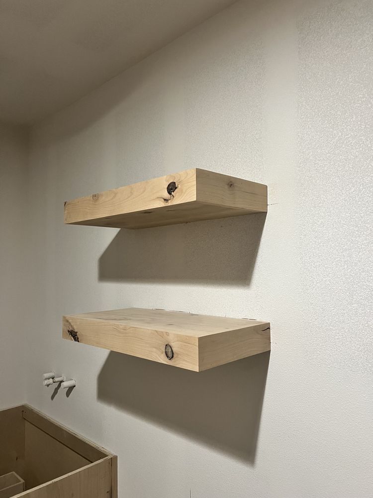 Two unfinished wooden shelves mounted on a white wall, with a cardboard box in the lower left corner