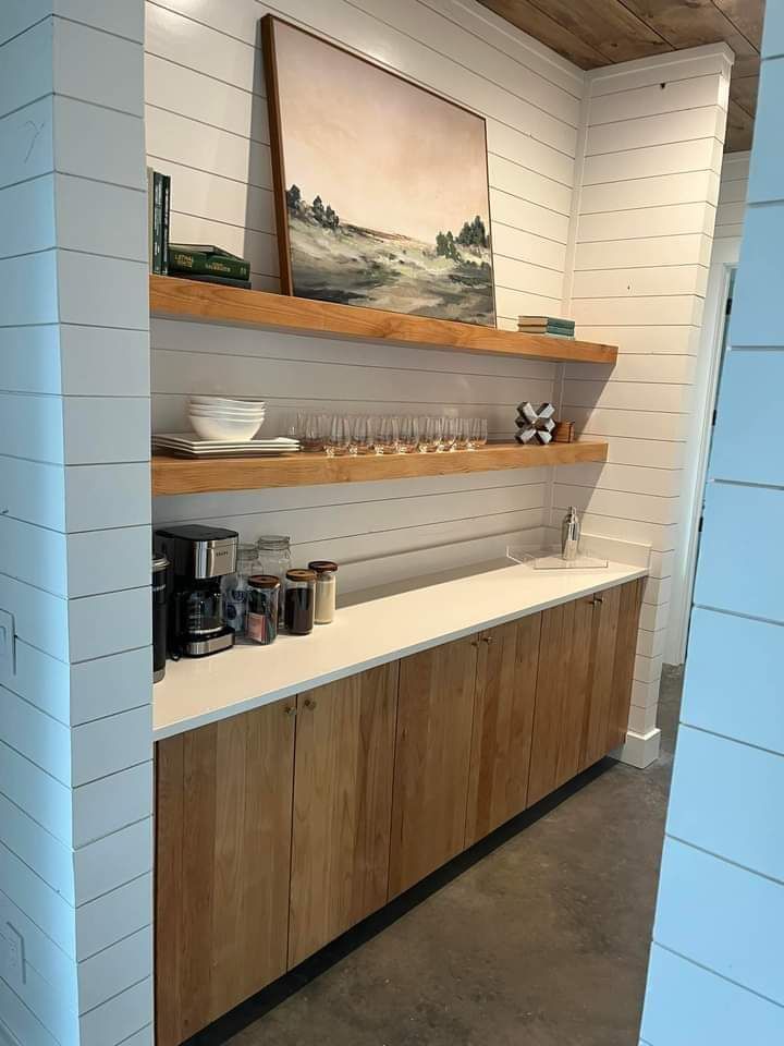 Coffee station with wooden shelves, cups, and a framed landscape painting in a white-paneled corner