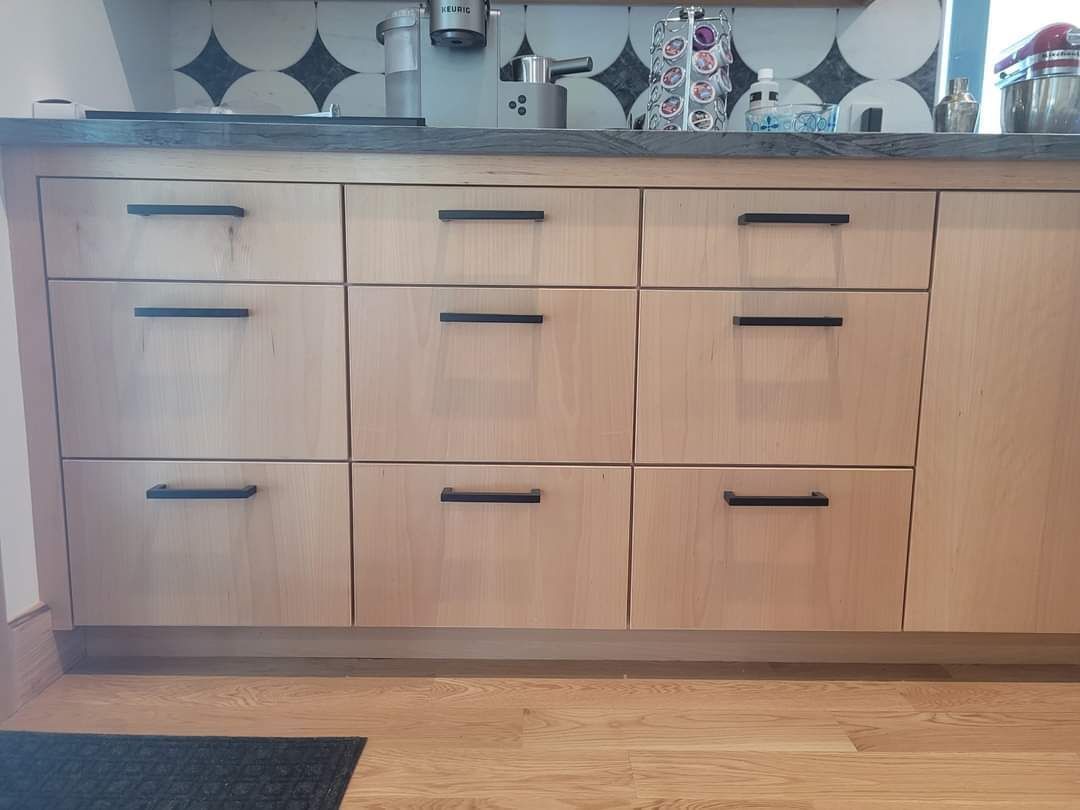 Light wood kitchen cabinet with black handles and countertop decor against a tiled backsplash.