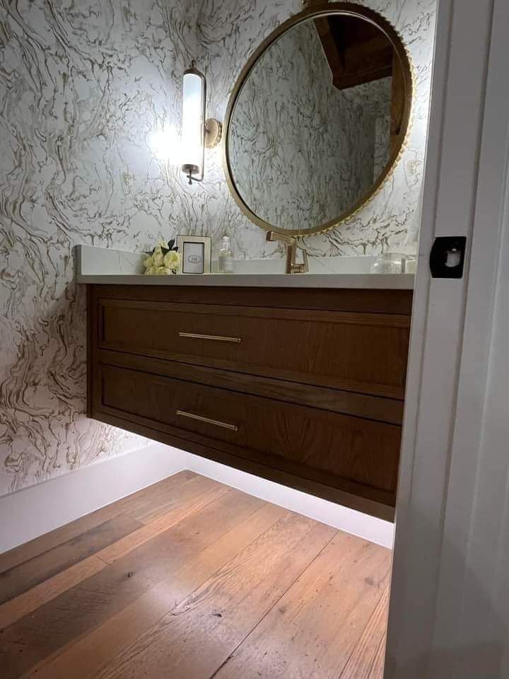 Elegant bathroom vanity with round mirror, wall sconce, marble-pattern wallpaper, and wooden floorboards.