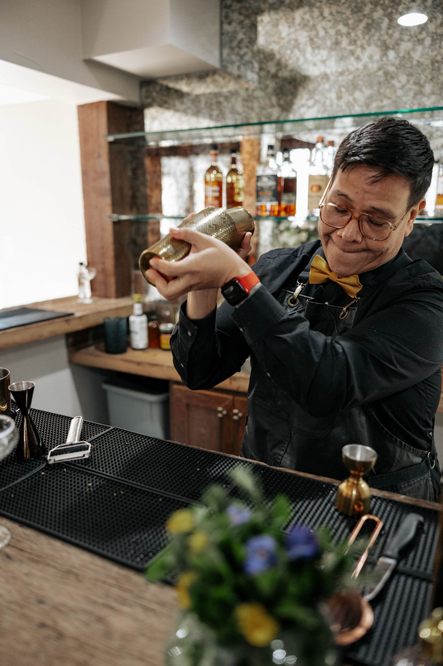 Bartender shaking a cocktail at a bar. He wears a yellow bow tie and vest, with a floral arrangement in foreground.