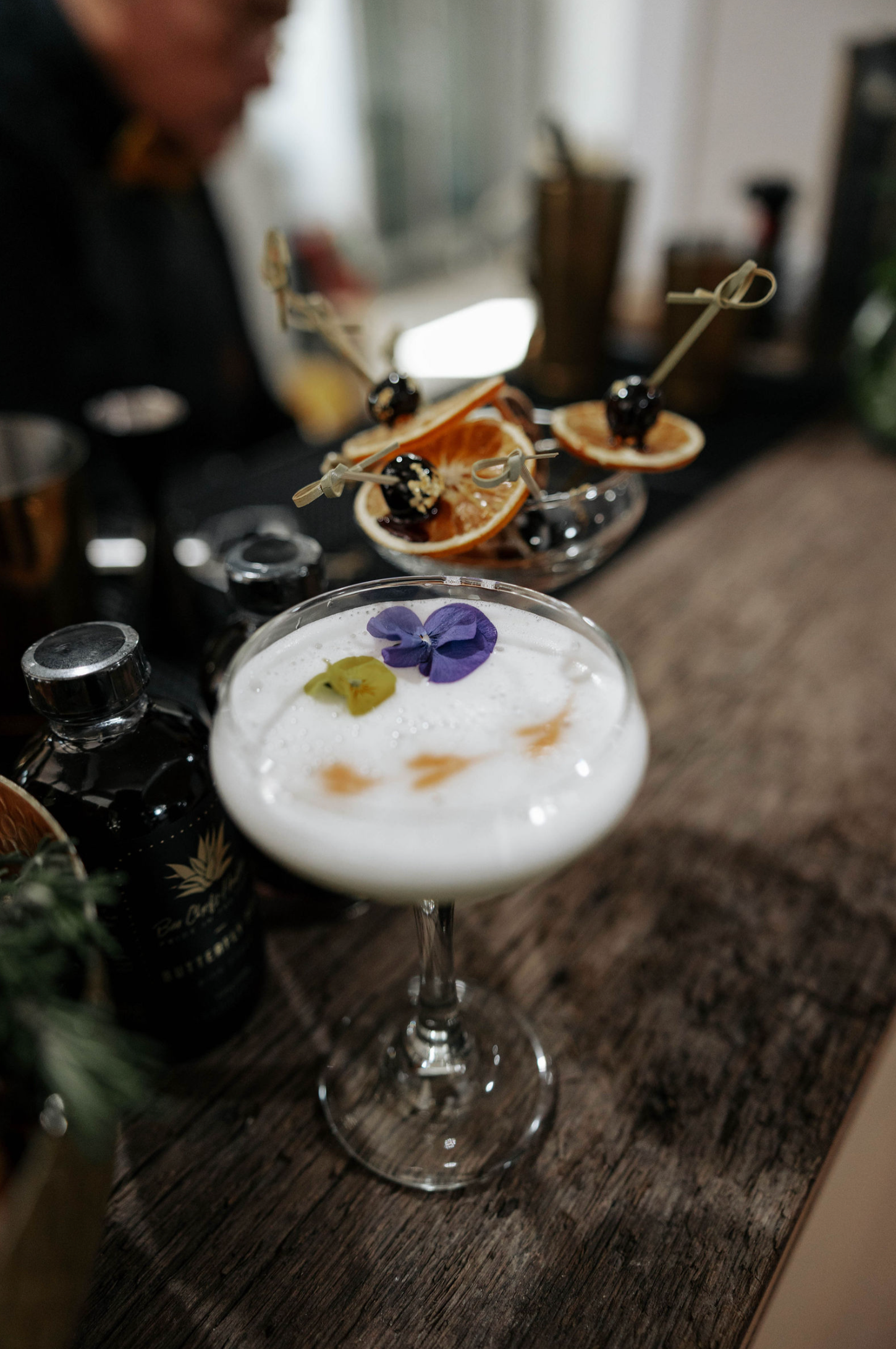 Cocktail in a coupe glass with foamy white top, garnished with flowers and orange slices, on a wooden surface.