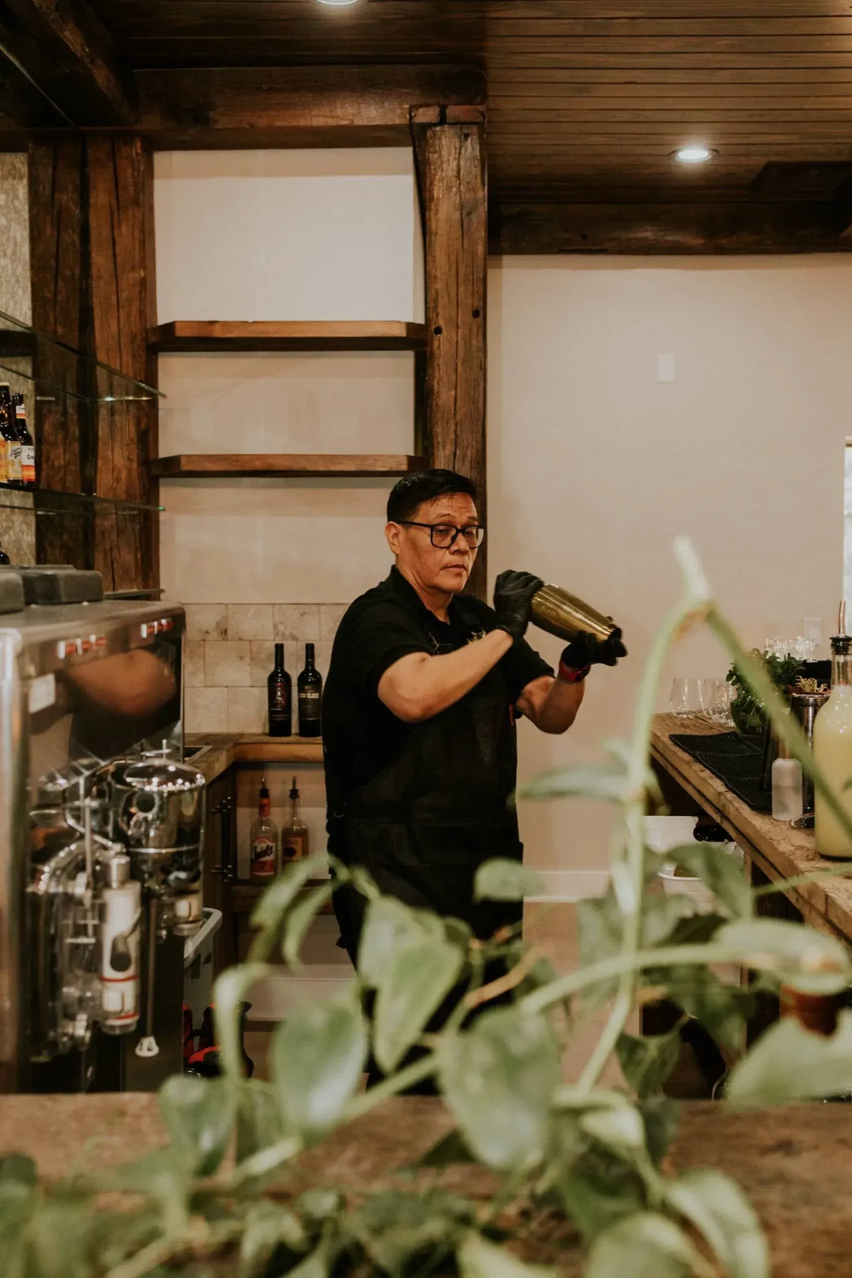 Bartender shaking a cocktail shaker behind a bar; wooden beams and neutral background.