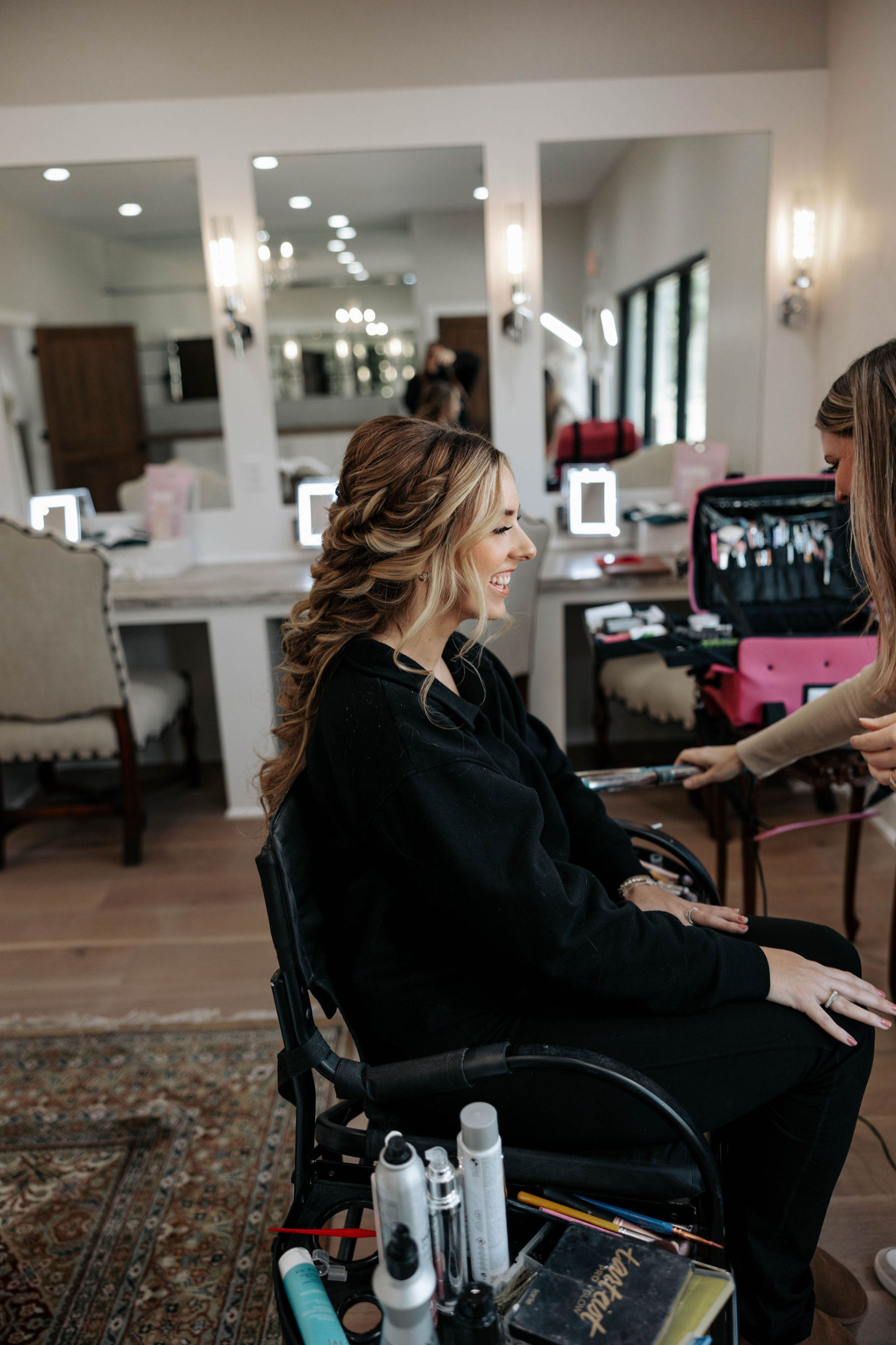 Woman in black outfit smiles while stylist works on her hair in a salon.