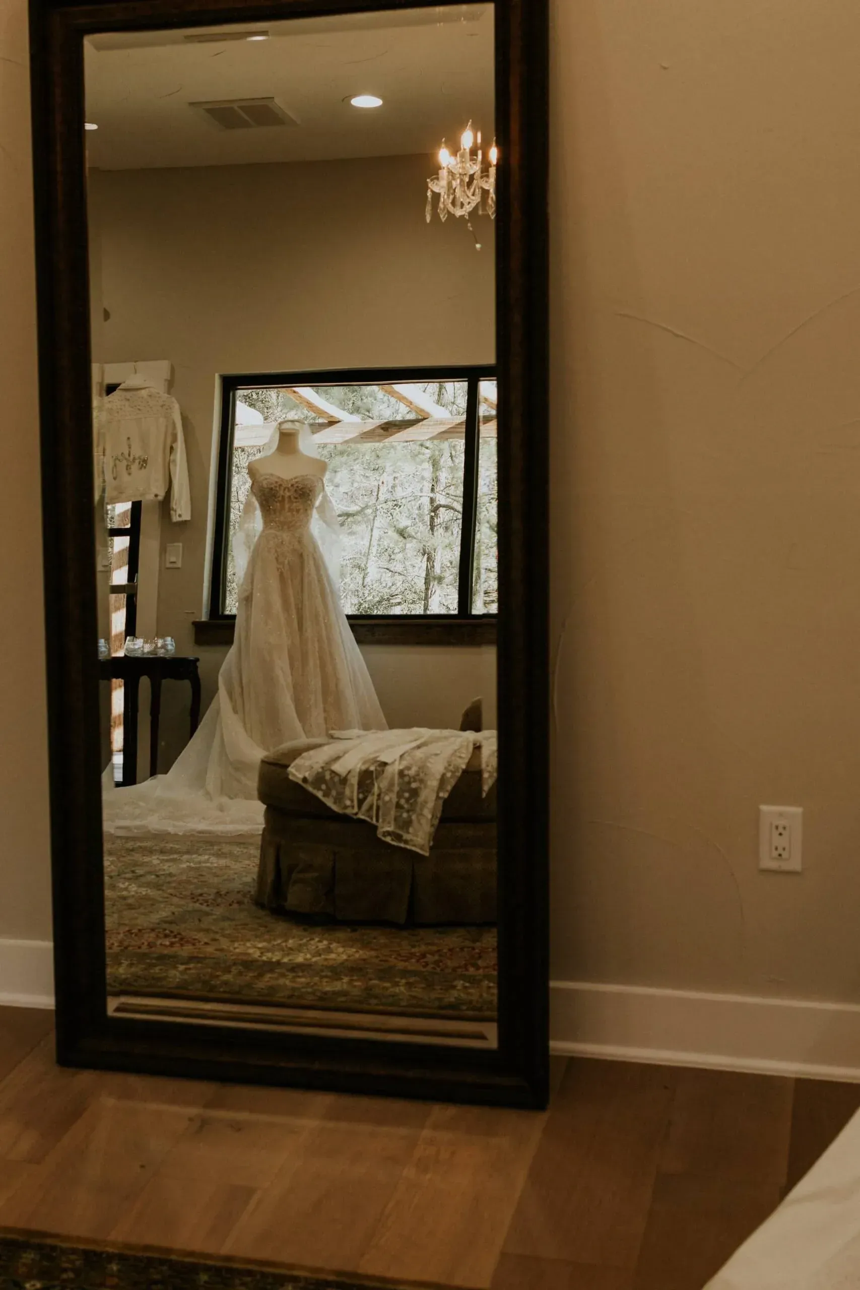 Full-length mirror reflecting a wedding dress on a mannequin in a room with a window and a chandelier.