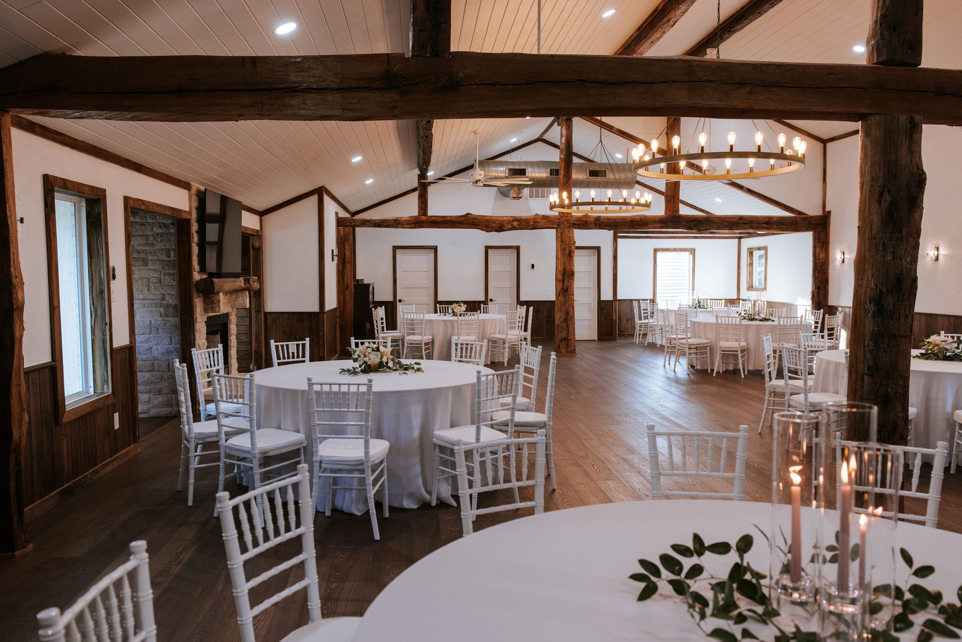 Wedding reception hall with round tables, white chairs, and wooden beams.