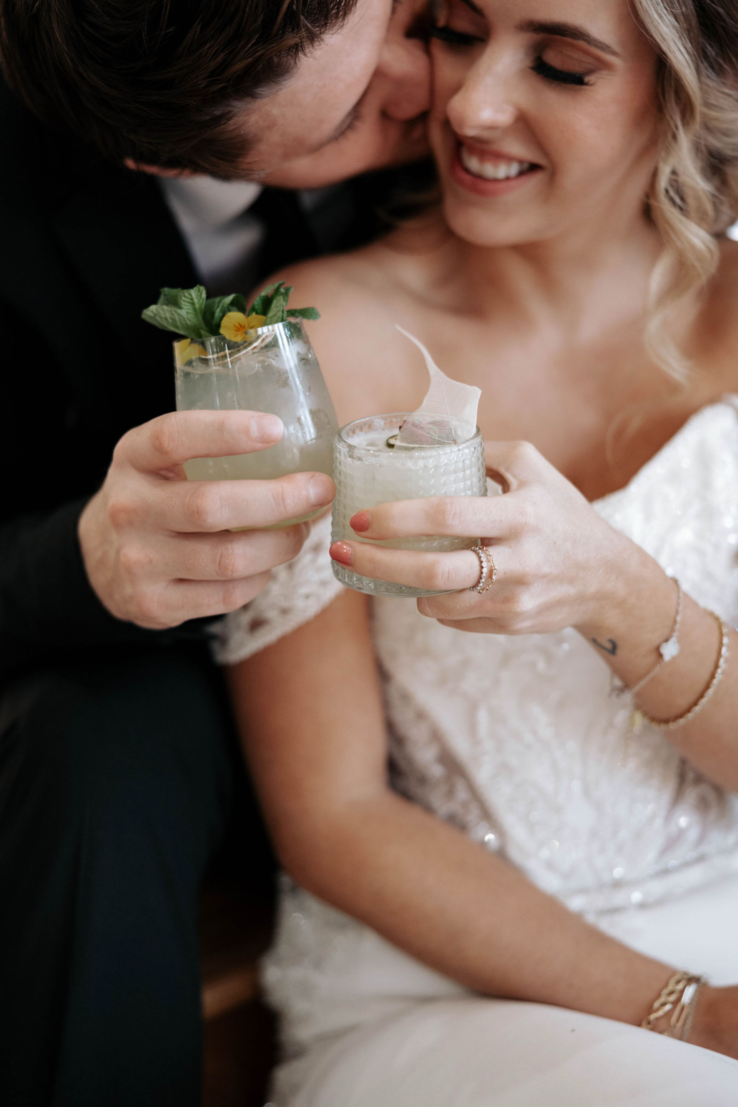 Couple toasting cocktails, man kisses woman's cheek, wearing wedding attire.