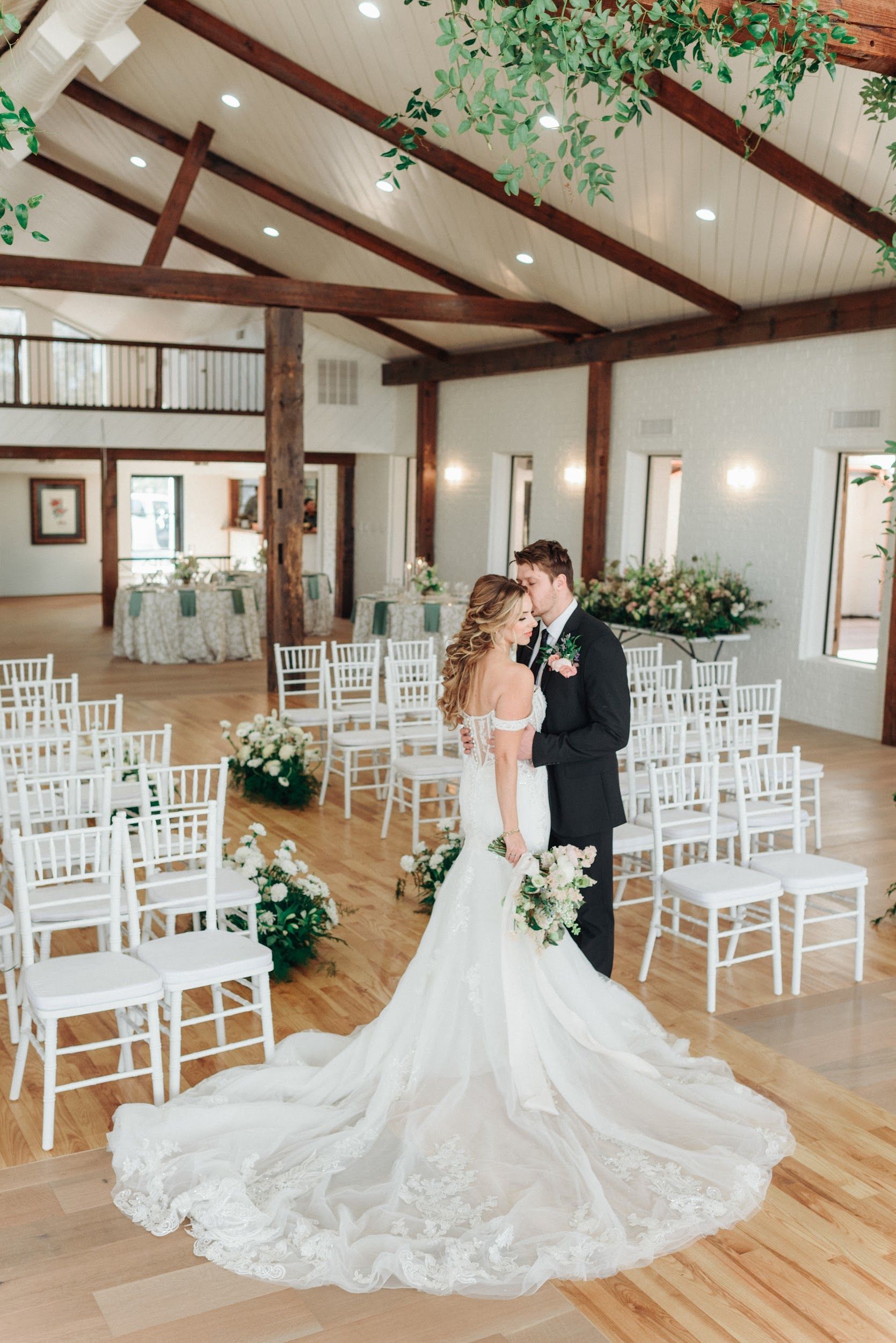 Bride and groom embracing in a light-filled wedding venue with rows of white chairs and floral arrangements.