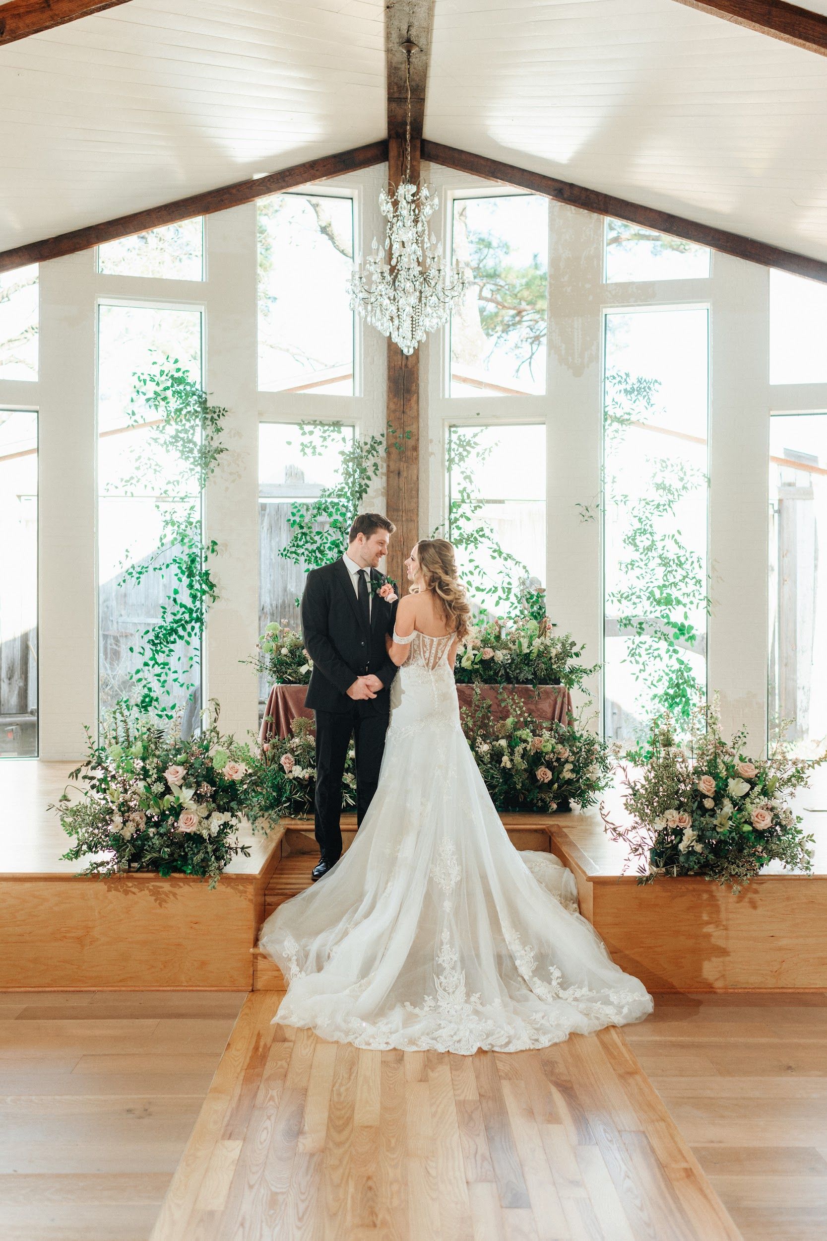Couple at altar in a bright room with flowers. Bride in white dress, groom in black suit.