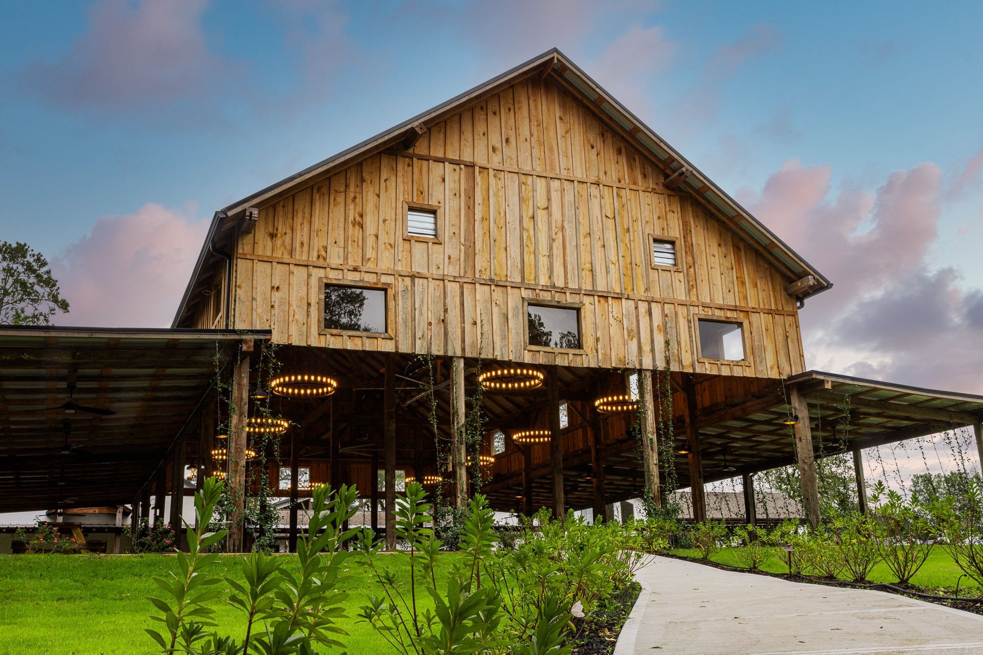 Rustic wooden barn with overhead patio, green grass, and pathway leading to the entrance under a sunset sky.