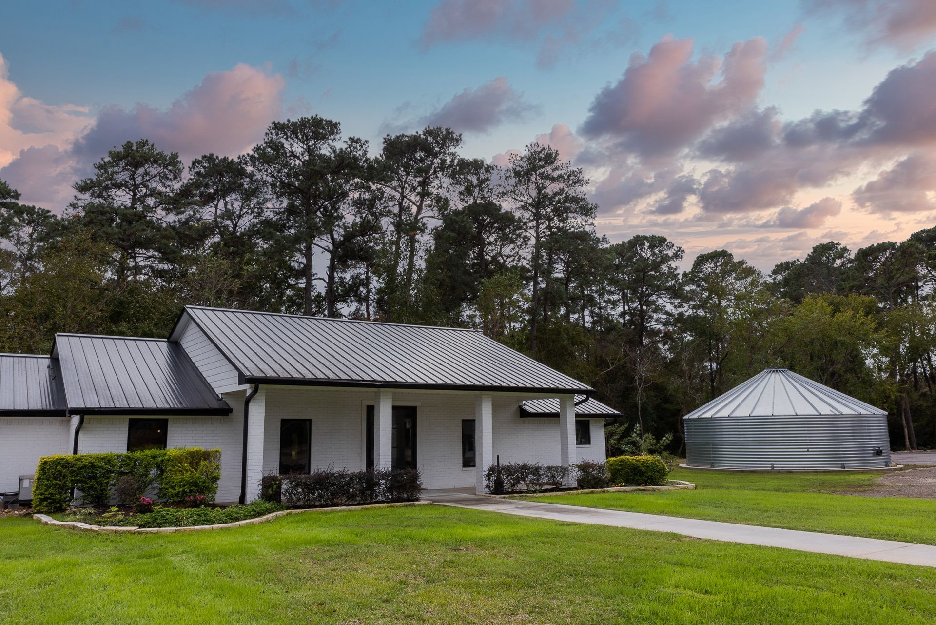 White house with silver roof and silo on a green lawn with a sunset sky and trees.