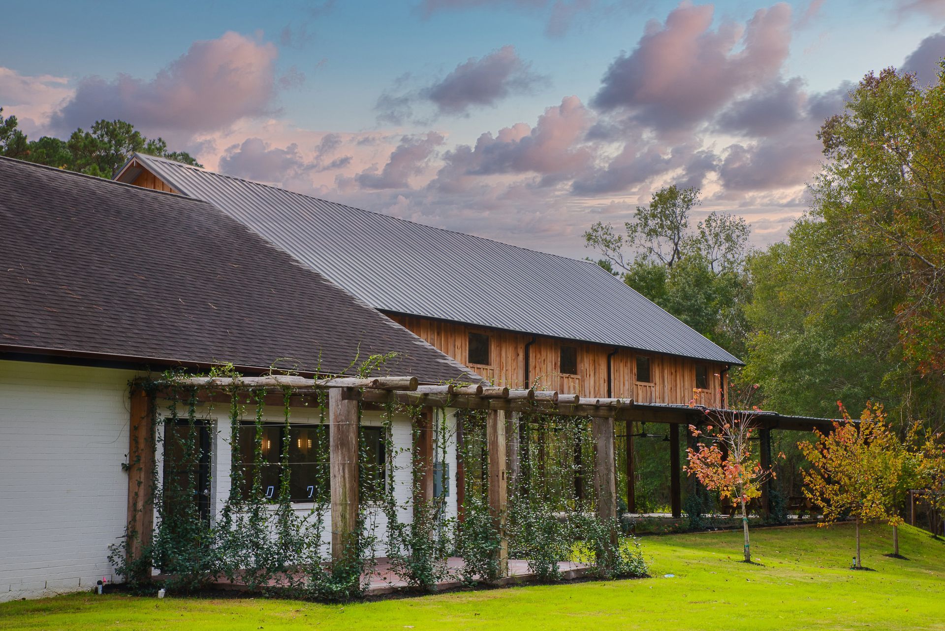 Barn-style building with a corrugated metal roof and a wooden pergola covered in vines, set against a cloudy sky.