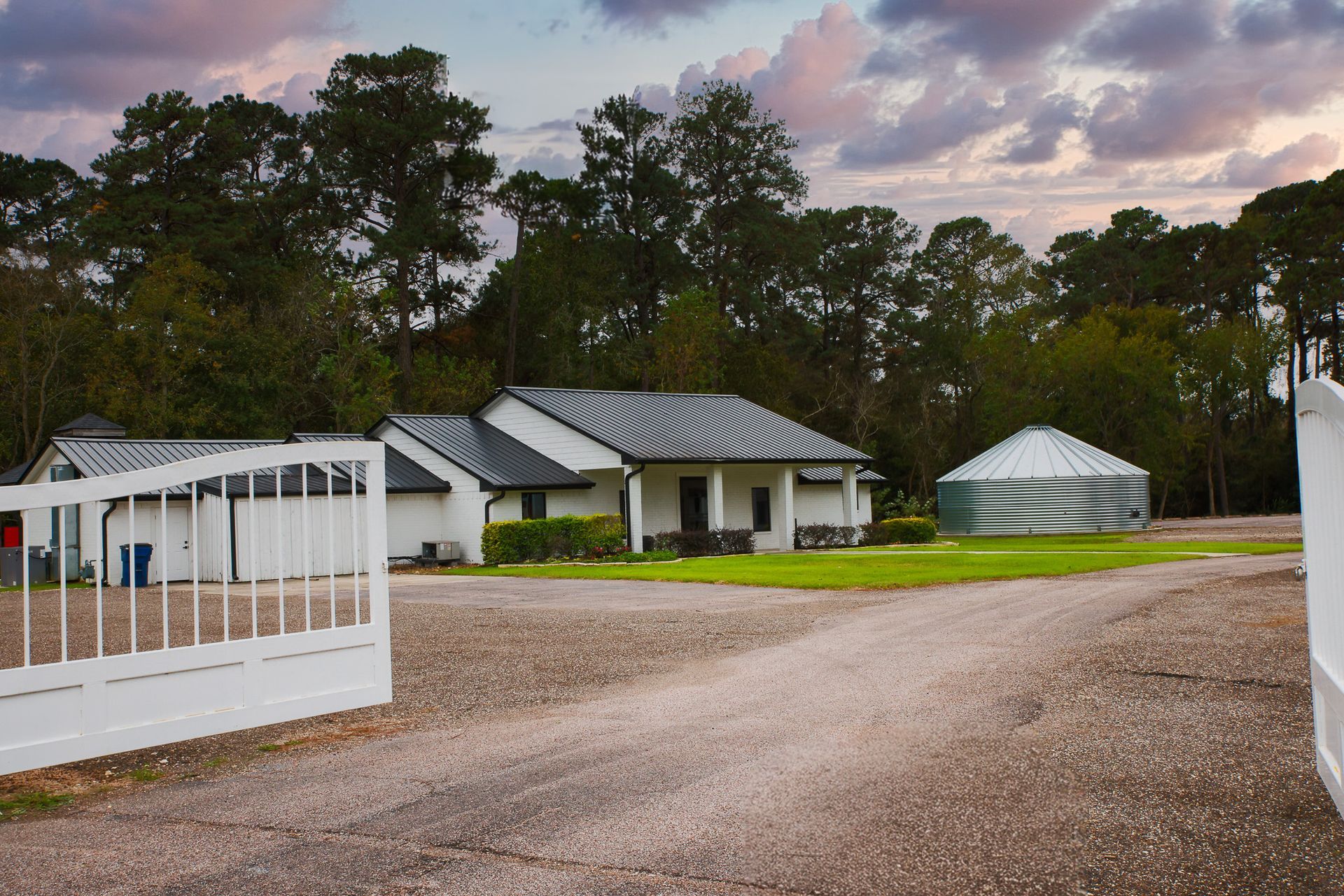White house with a metal roof behind a white gate on a gravel driveway, with trees in the background.